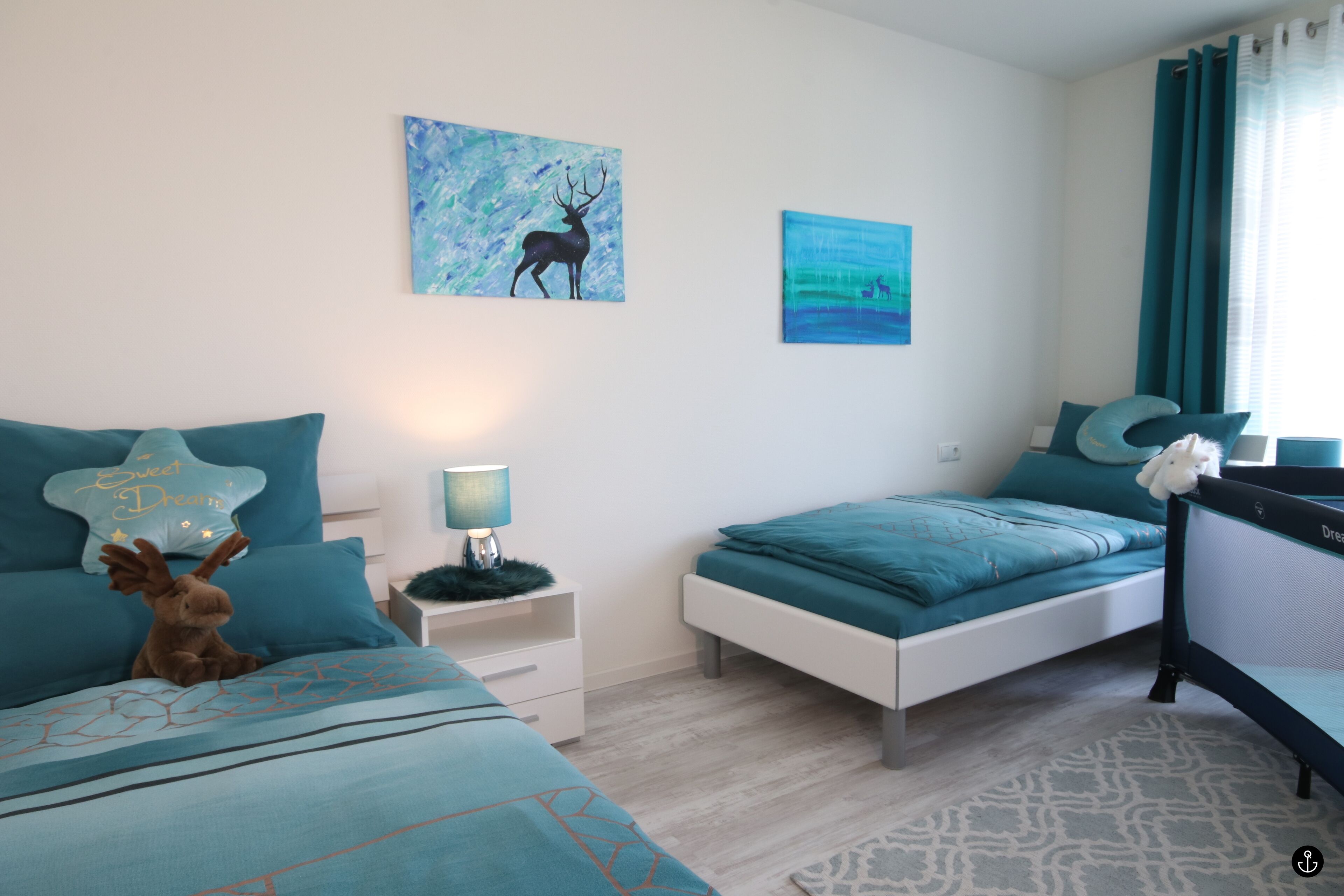 A bedroom with two beds, teal bedding, a star-shaped pillow, a moose plush toy, and two paintings on the wall.