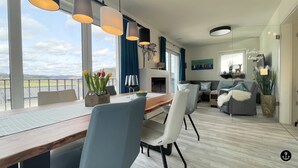 A modern living room with a wooden dining table, white chairs, and a grey sofa. There is a fireplace, a large window with a view, and a painting on the wall. - Sonnenweg 36 - LAKEVIEW HOUSE TÜRKIS (Diemelsee)