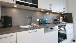 A modern kitchen with white cabinets, a stainless steel sink, a black coffee maker, a blue toaster, and a built-in oven. - Sonnenweg 36 - LAKEVIEW HOUSE TÜRKIS (Diemelsee)