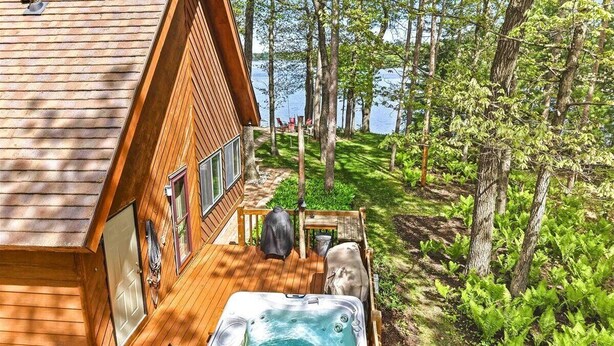 En stuga byggd av trä med ett bubbelbad på en veranda, omgiven av träd och en sjö.