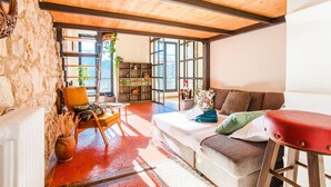 A cozy living room with a wooden ceiling, a stone wall, a red floor, a sofa with cushions, a red stool, and a bookshelf.