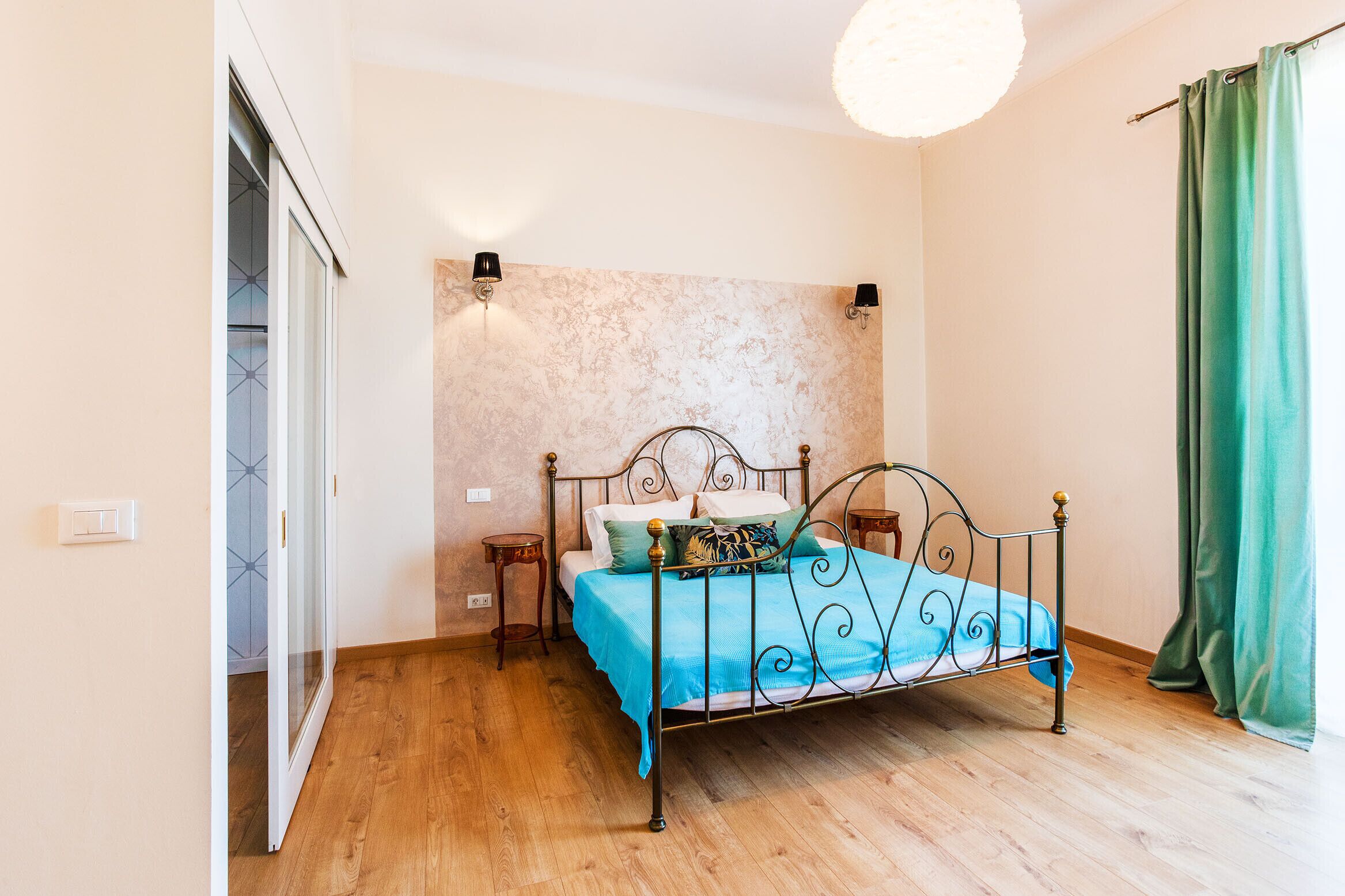 A bedroom with a metal bed frame, a blue bedspread, two bedside tables, and a wall-mounted light fixture.