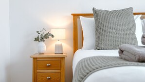 Un lit bien fait, avec un oreiller à motifs gris et un oreiller blanc, une lampe de chevet et une table de chevet en bois sur laquelle est posé un vase rempli de verdure.
