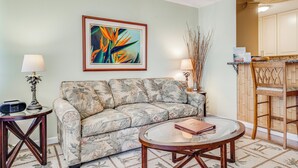 A living room with a sofa, a coffee table, a framed painting, a lamp, and a kitchen area with cabinets.