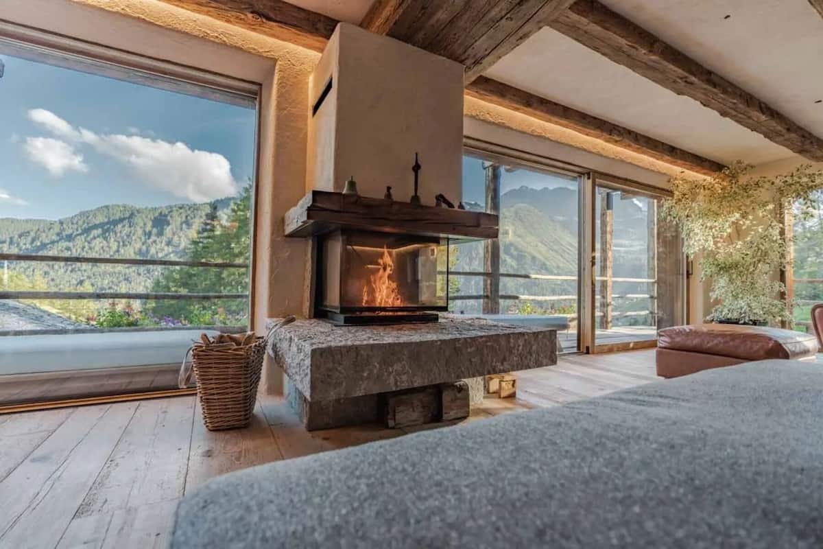 Una habitación con chimenea de piedra, ventanas amplias con vistas a la montaña y vigas de madera en el techo.