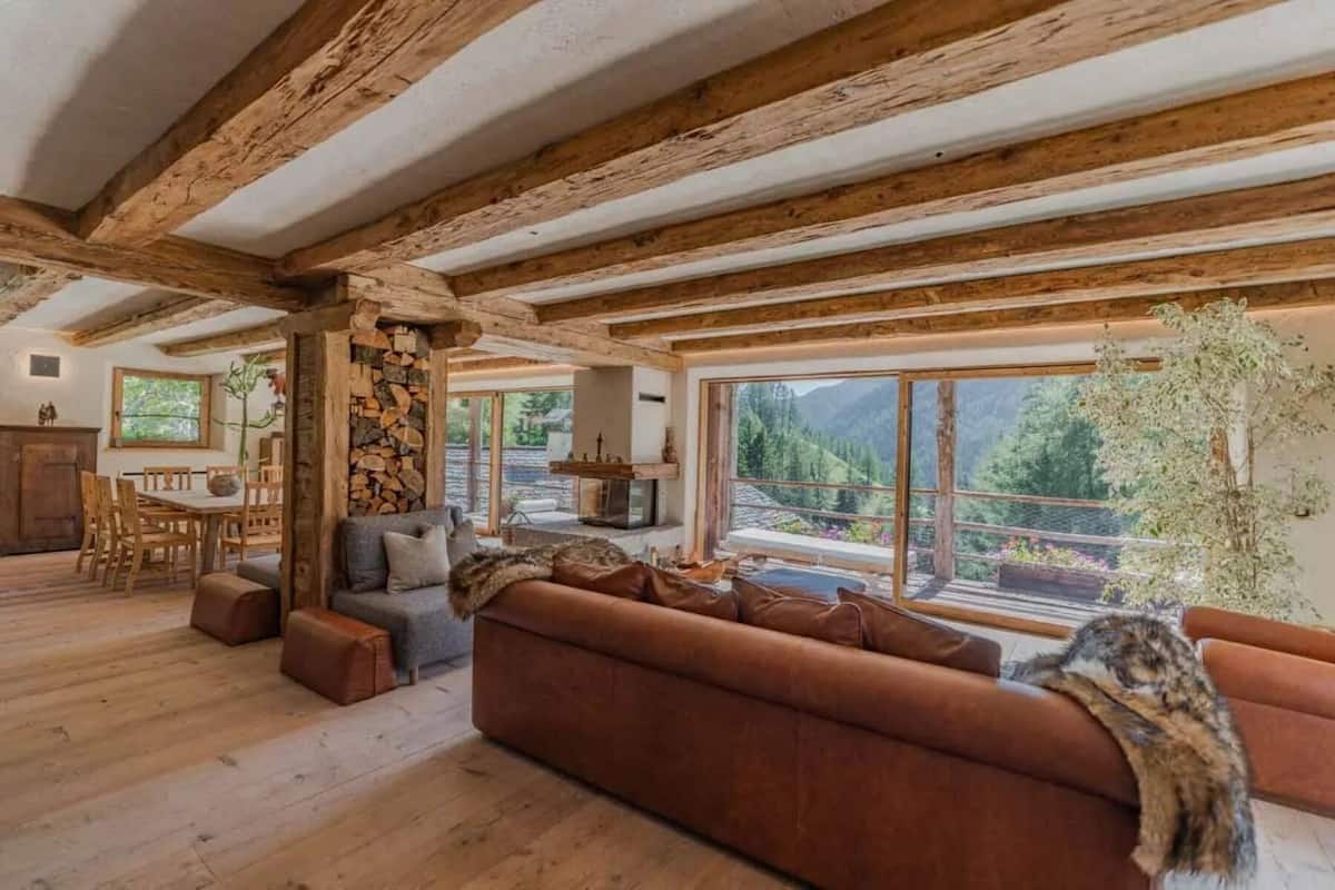 Un interior rústico con vigas de madera a la vista, chimenea de piedra, sofá y vista a un paisaje montañoso.
