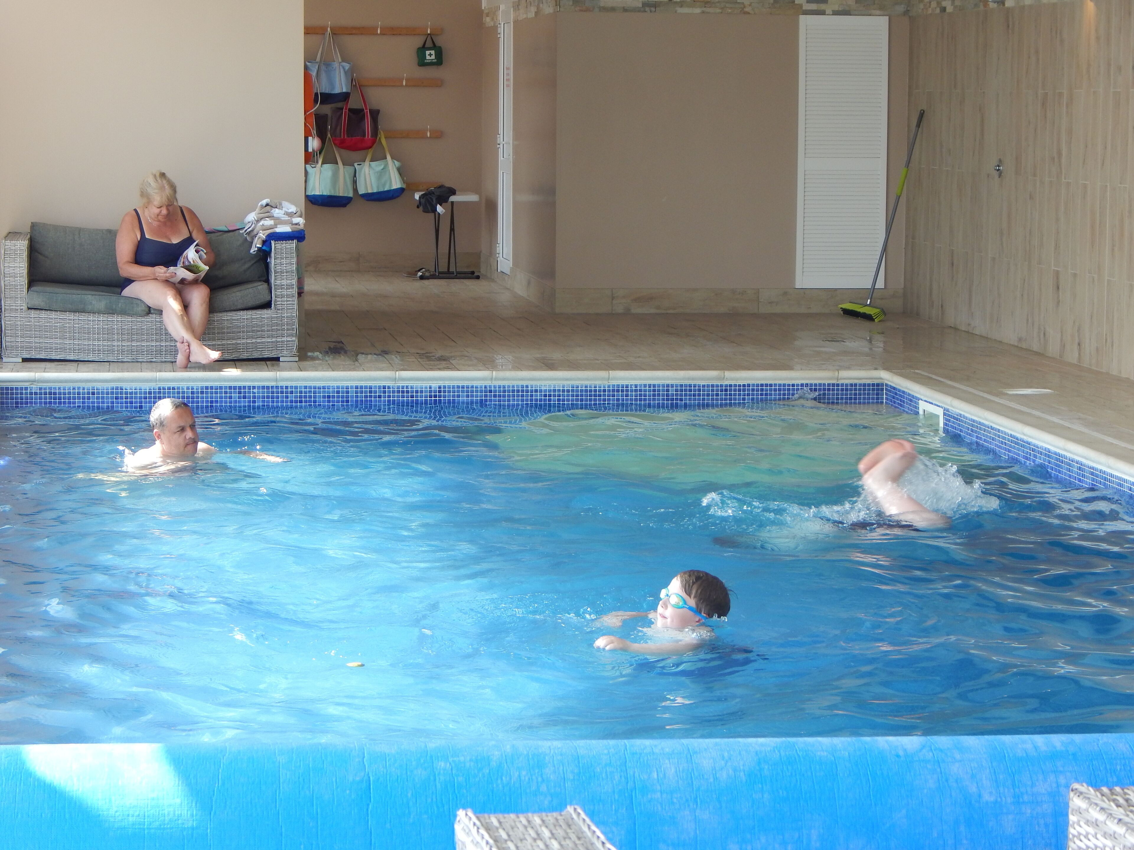 A swimming pool with three people in the water, a lounge area with a sofa and a table, and a tiled wall with hanging bags.