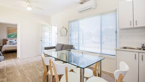 A modern, well-lit apartment with a glass dining table, white chairs, a grey sofa, and a kitchen area with white cabinets and a sink.