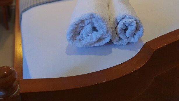 En snyggt bäddad säng med vita lakan och två hoprullade handdukar på sängen.