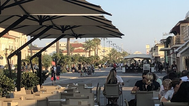 Un café en plein air avec des tables et des chaises sous des parasols, une rue en arrière-plan et des bâtiments de chaque côté.