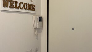 Une pièce avec une porte fermée, un interphone fixé au mur, une pancarte « BIENVENUE » et un thermostat.