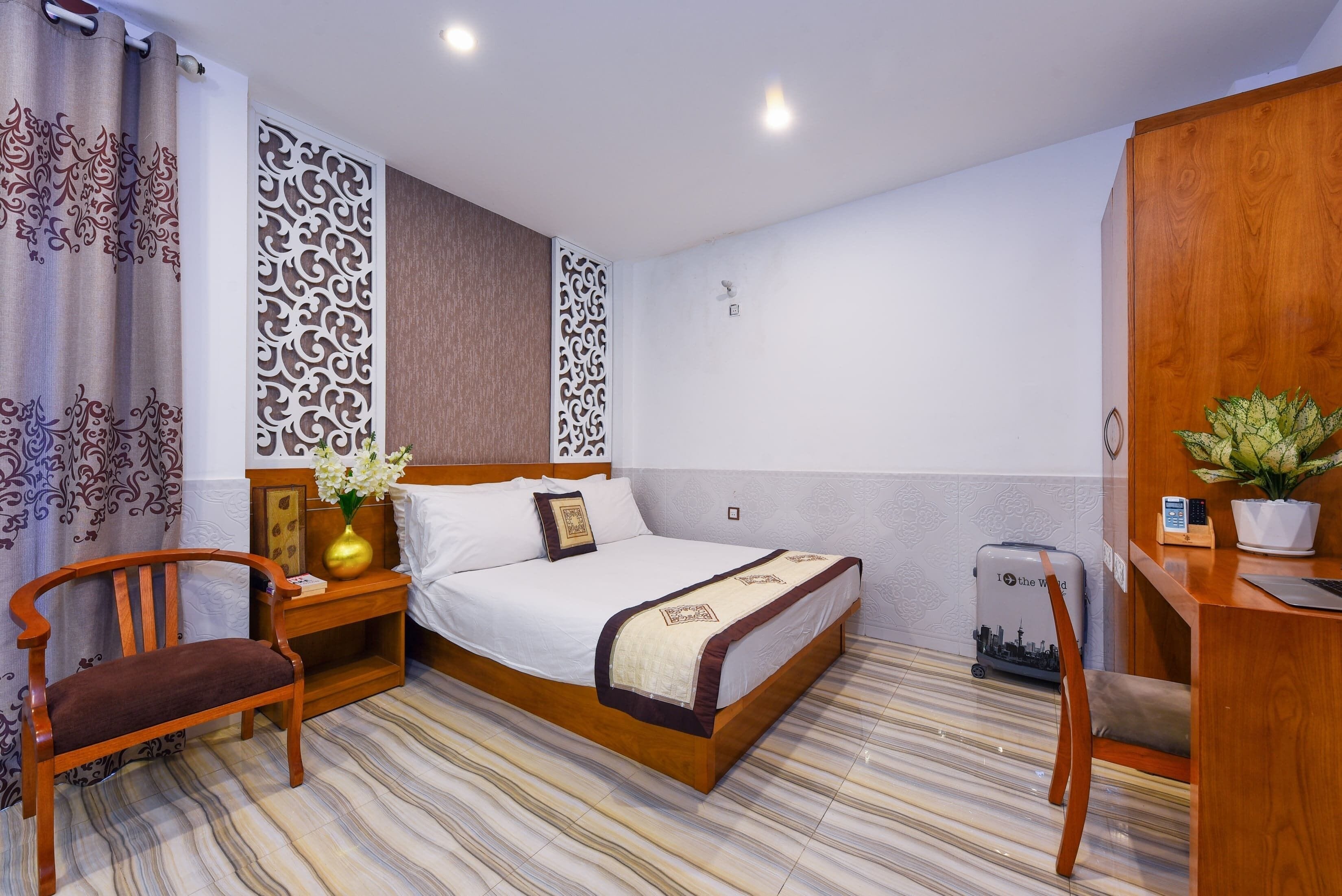 A hotel room with a wooden bed, a wooden chair, a desk with a plant, and a suitcase.