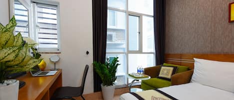 A hotel room with a bed, a desk with a laptop, a chair, a small table with a plant, and a view of the outside through large windows.