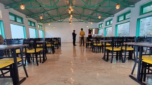 A spacious indoor dining area with high ceilings, green structural beams, and large windows. Tables and chairs are arranged neatly, and the floor is polished marble. - LaSerene The Cottage (Nainital)