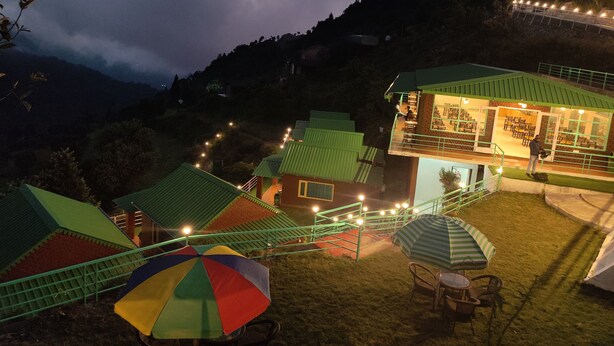 A hillside accommodation with multiple green-roofed buildings, outdoor seating, and a dining area visible through the windows. - LaSerene The Cottage (Nainital)