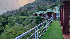 A hillside with several small, single-story buildings with green roofs, surrounded by lush greenery and a white metal railing. - LaSerene The Cottage (Nainital)