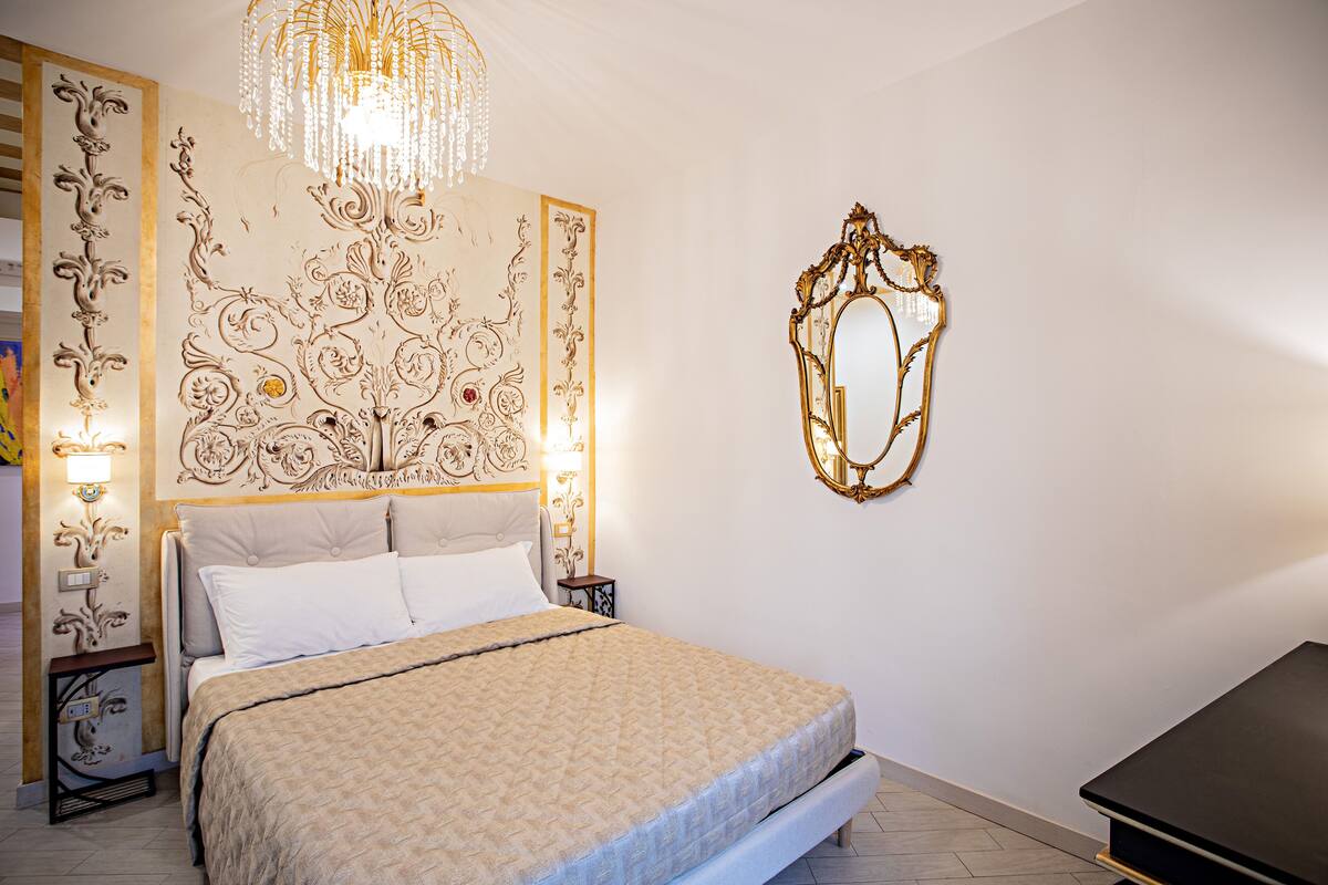 A neatly arranged bedroom with a large bed, a chandelier, and a decorative mirror on the wall.