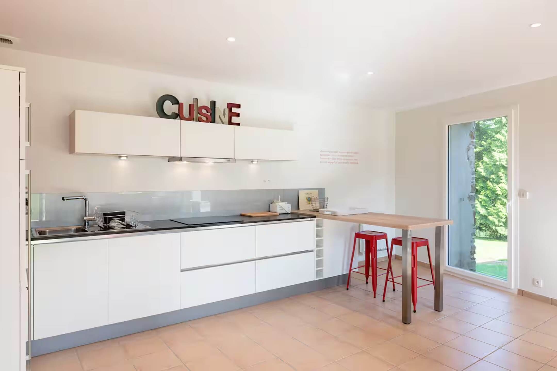 Une cuisine moderne avec des meubles blancs, un plan de travail noir et un îlot central en bois. On y trouve deux tabourets rouges et une fenêtre donnant sur un espace verdoyant.