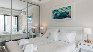 A modern hotel room with a large bed, white linens, a bedside table with a lamp, a wall-mounted painting, and a large mirror reflecting the room. - Liberty's Canvas - Balcony Suite near Acropolis (Αθήνα)