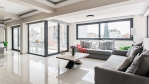 A modern living room with a large window, a grey sofa, a coffee table, and a dining area visible through the glass doors. - Liberty's Canvas - Balcony Suite near Acropolis (Αθήνα)