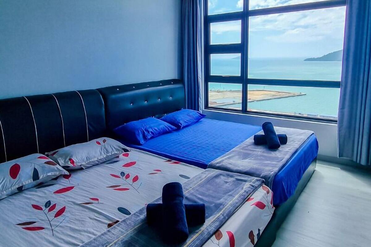A hotel room with two beds, a large window with a view of the sea, and a blue bedspread.