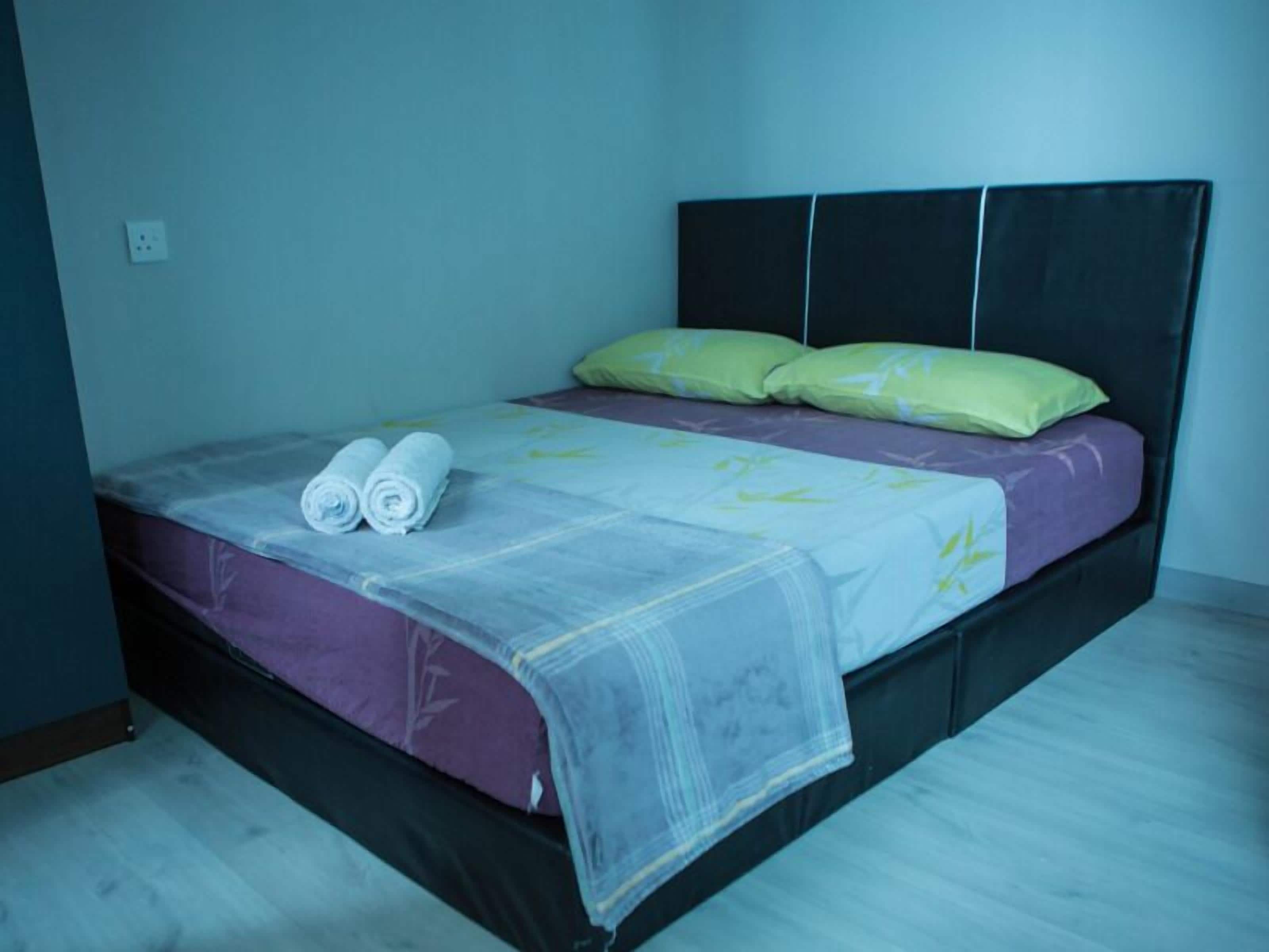 A single bed with a black headboard, a purple mattress, and two green pillows. There are two rolled towels on the bed.