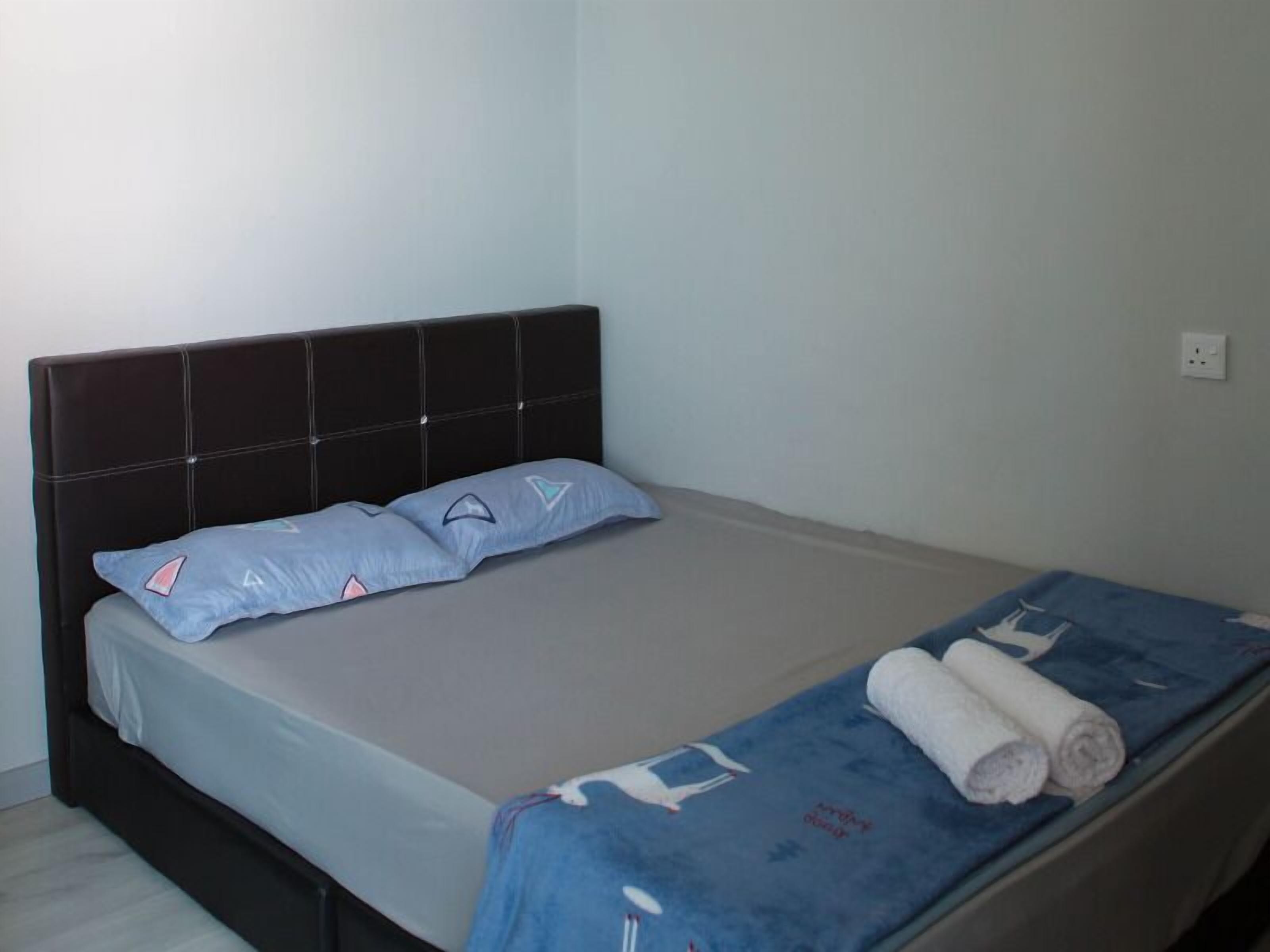 A bed with a dark headboard, two pillows, a blue blanket with a pattern, and a rolled white towel.