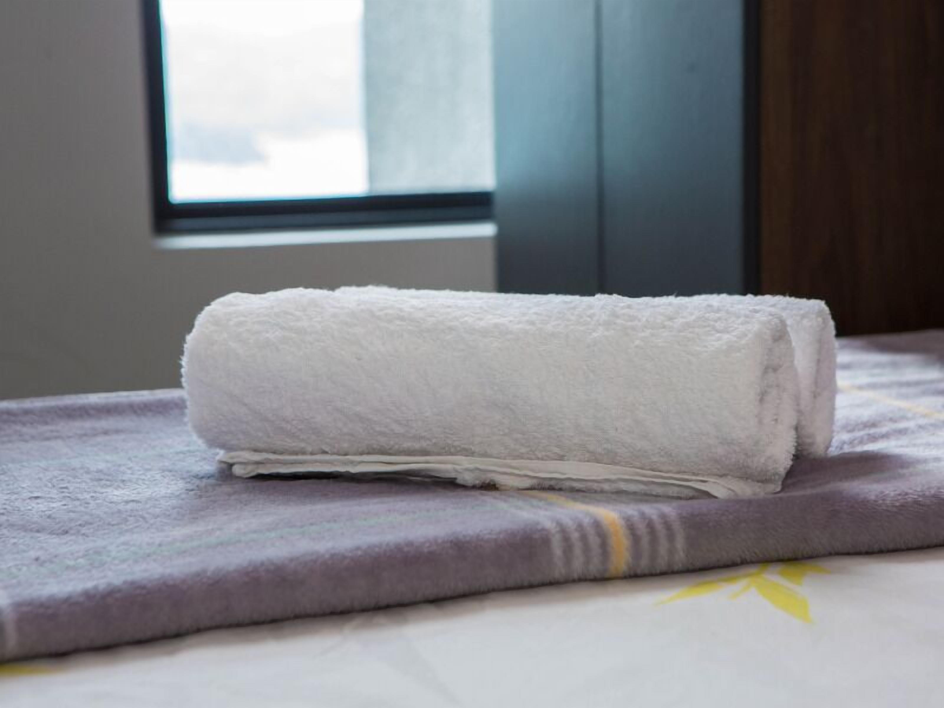 Toalha branca dobrada sobre uma cama com manta roxa e padrão floral.