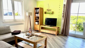 En opholdsstue med sofa, sofabord, fjernsyn på et træstel, reol og et vindue med gardiner.