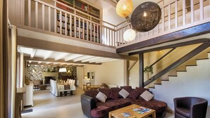 Un vaste espace de vie comprenant un grand canapé d’angle, une table à manger et un escalier menant à un niveau supérieur où se trouvent des étagères à livres.