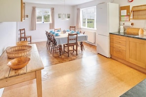 A dining area with a wooden table and chairs, a kitchen with wooden cabinets, and a view of the outdoors through large windows.