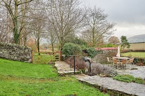 A stone pathway leading to a rustic outdoor seating area with a table and chairs, surrounded by greenery and a stone wall.