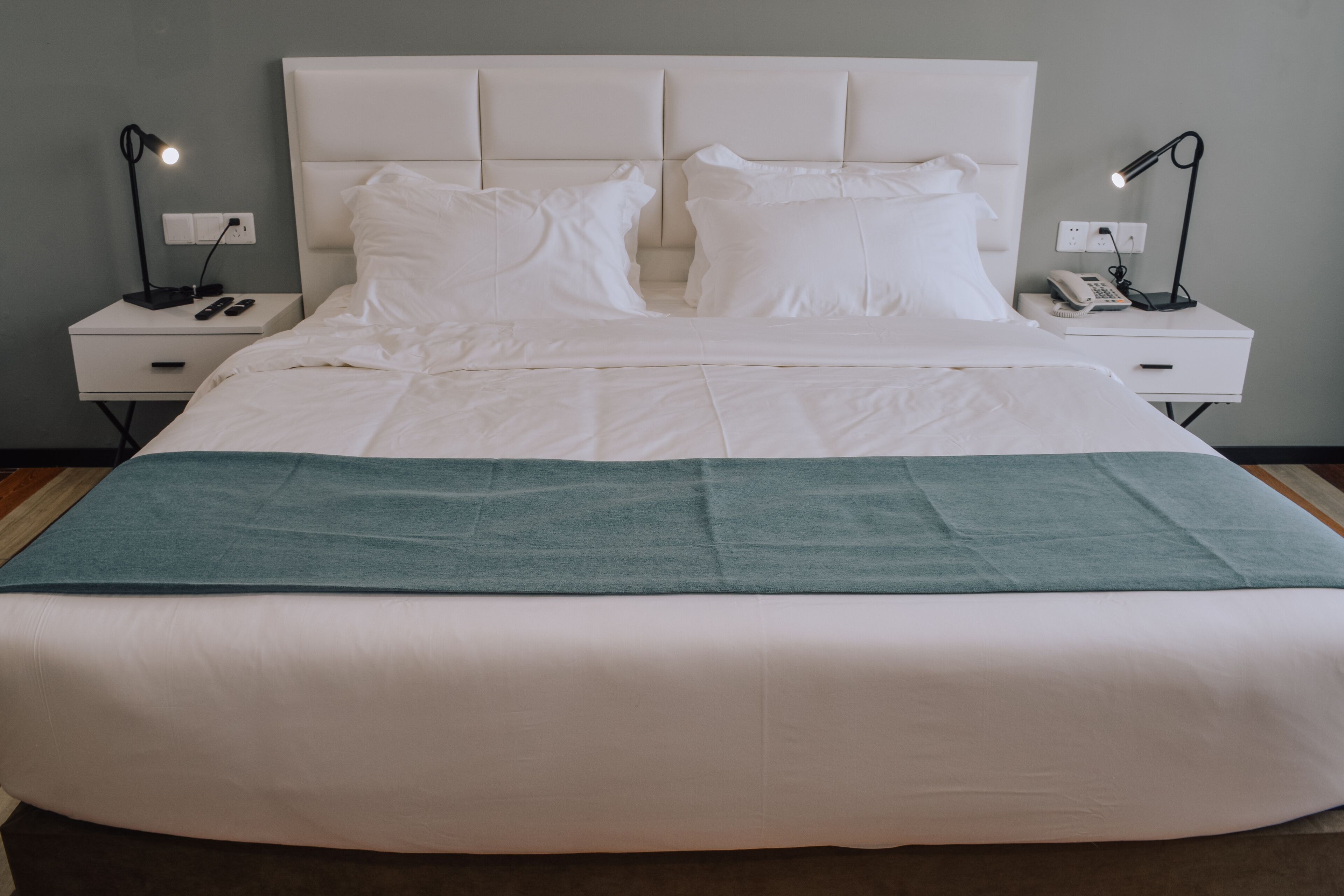A neatly made bed with white linens and a teal throw blanket, flanked by two bedside tables with lamps and a phone.