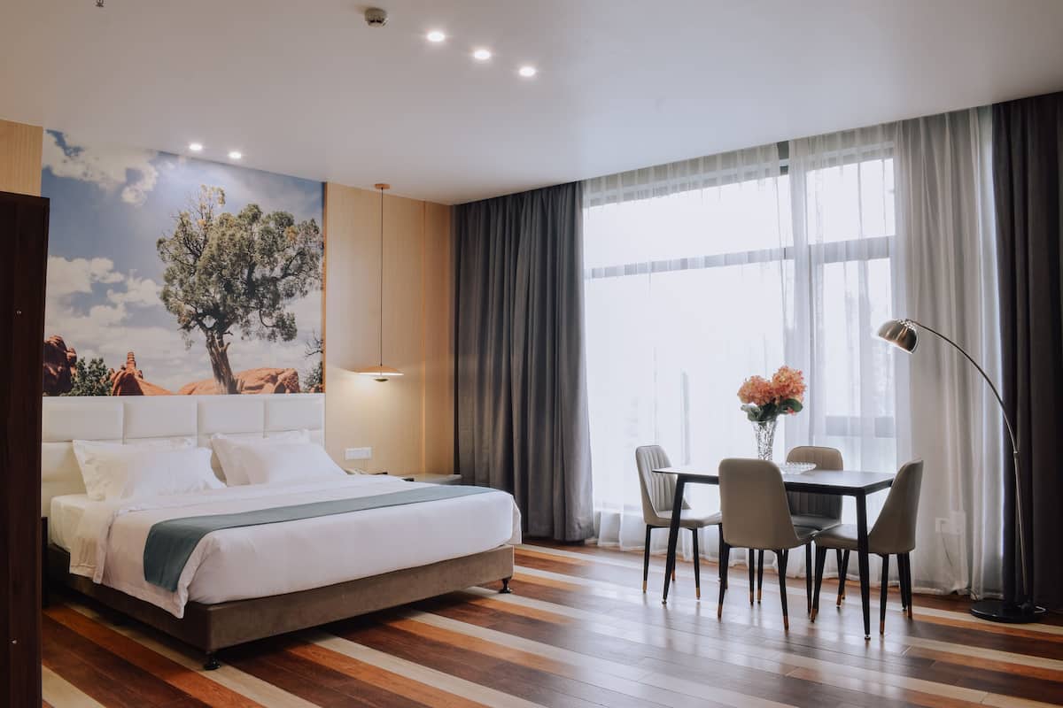 A hotel room with a bed, a dining table with chairs, a lamp, and a large window with curtains.