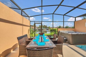 Um pátio externo com mesa posta para quatro pessoas, cercado por cadeiras de vime, uma banheira de hidromassagem e vista para uma piscina e vegetação.