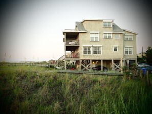 A two-story house with a balcony on the upper floor, situated on stilts over a grassy area.