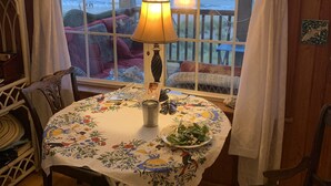Habitació acollidora amb interior de fusta, taula rodona amb mantell de flors, una làmpada i vista a la platja a través d'una finestra gran amb cortines blanques.