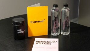 Eine Handdesinfektionsflasche, eine gelbe Karte mit dem Schriftzug 'wynnwood', zwei Wasserflaschen und ein Willkommensgruß liegen auf einem Tisch.