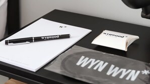 Una mesa negra con un bloc de notas blanco, un bolígrafo negro y un cuadernillo pequeño con la inscripción 'WYN WYN'.