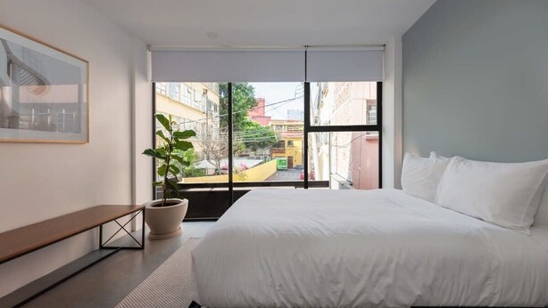 Un dormitorio moderno con una cama grande, una planta en maceta, un banco y vistas de la ciudad a través de ventanales.