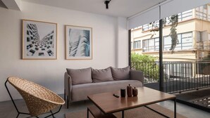 Una sala de estar moderna con un sofá, una mesa de centro y una silla de mimbre. Hay dos obras de arte enmarcadas en la pared.