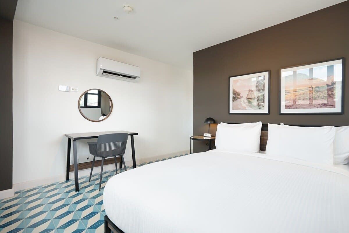 A hotel room with a large bed, a desk with a chair, an air conditioning unit, a round mirror, and two framed pictures on the wall.