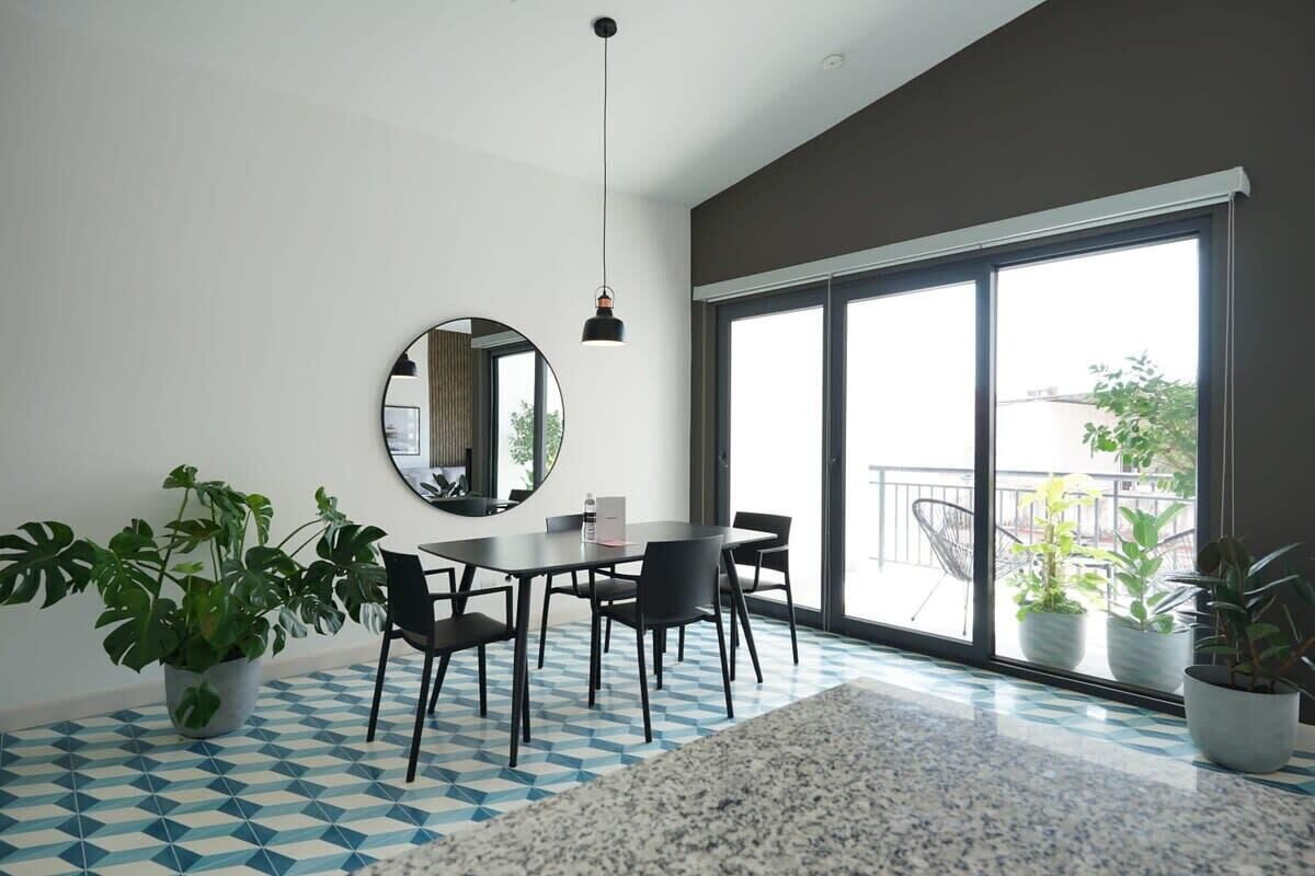 A modern dining area with a round table, black chairs, a hanging pendant light, and large windows with a view of the outdoors.