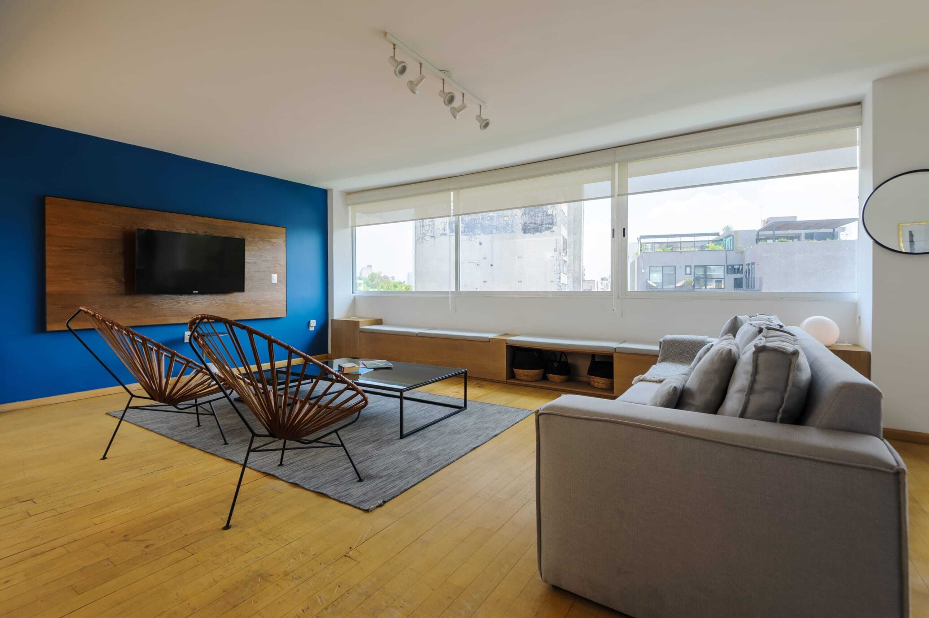 A modern living room with a large window, a flat-screen TV mounted on a wooden wall, a grey sofa, and two metal chairs.