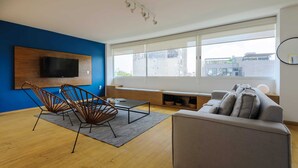 A modern living room with a large window, a flat-screen TV mounted on a wooden wall, a grey sofa, and two metal chairs. - w* | Polished 3BR Duplex in Condesa (Ciudad de México)