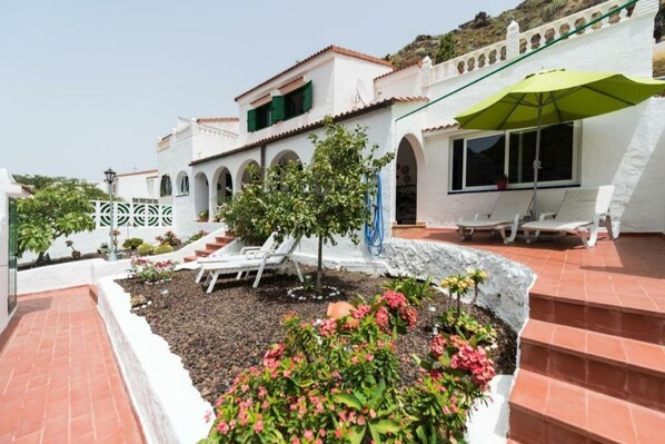 A white building with a tiled roof, a garden with flowers and a tree, and a patio with lounge chairs.