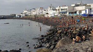 Een druk strand vol mensen die zonnebaden en zwemmen, omringd door witte gebouwen met balkons.