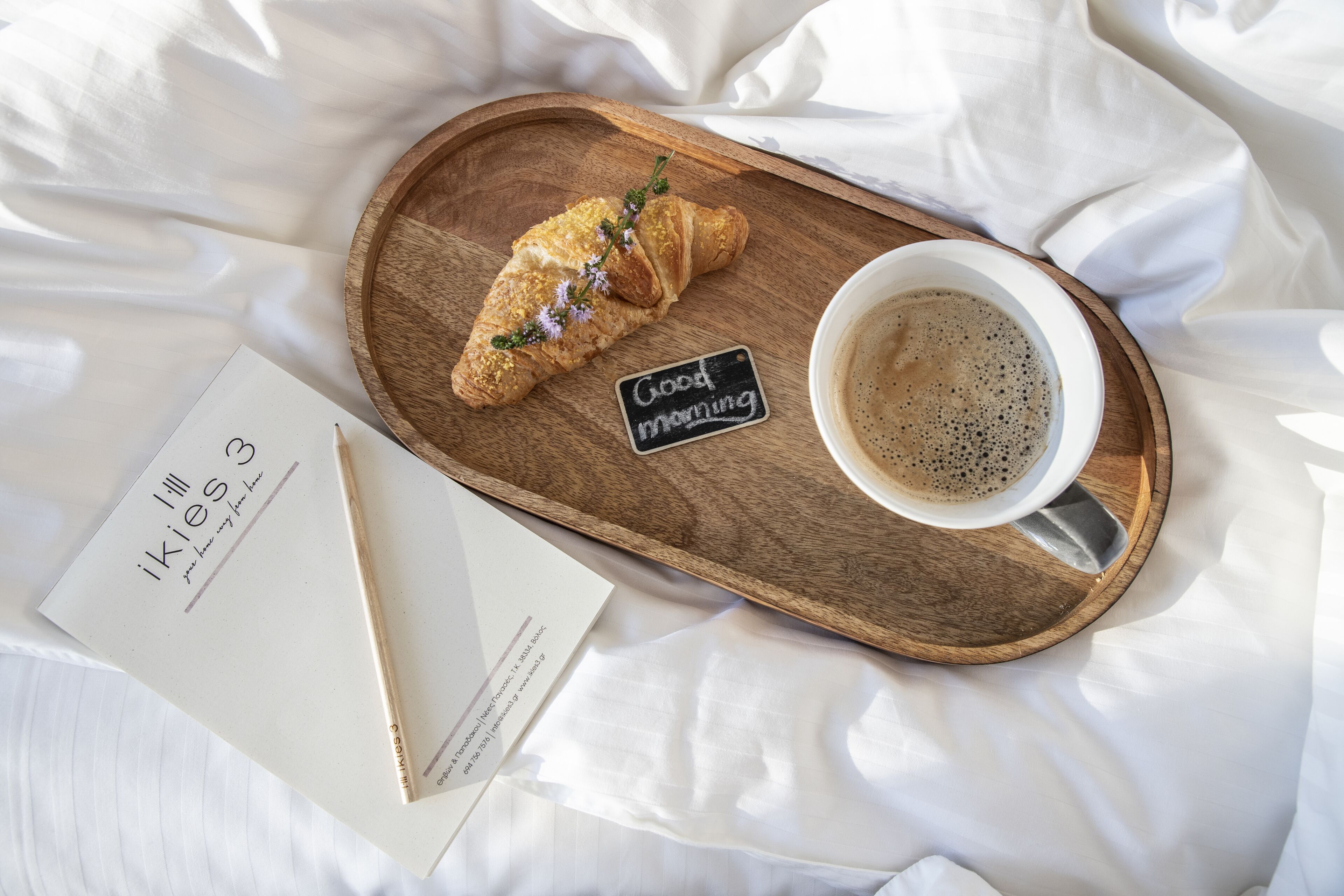 A wooden tray with a croissant and a cup of coffee on a bed with white sheets.