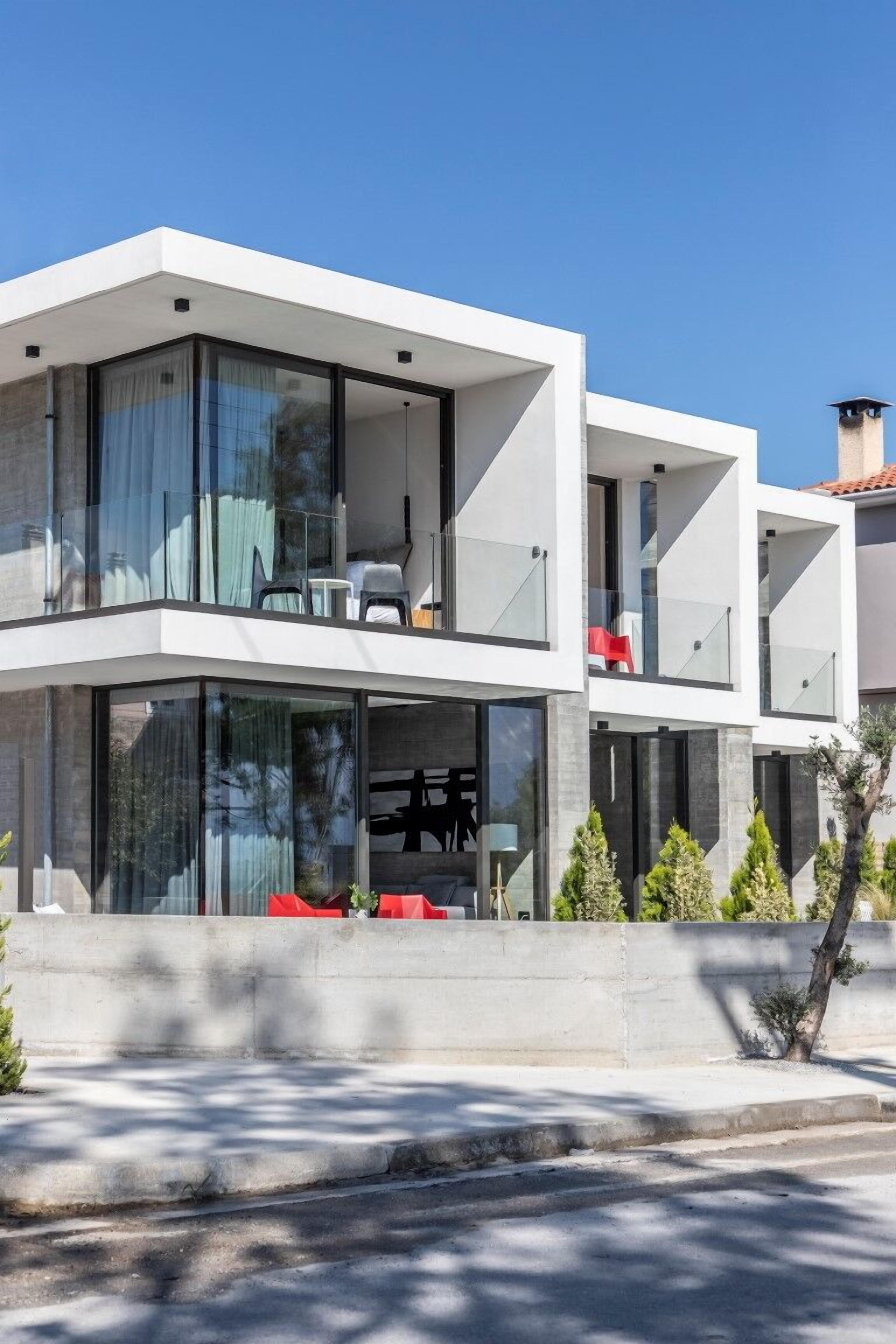 Een modern, meerlaags huis met grote glasramen, een balkon met rode stoelen en een betonnen fundering.