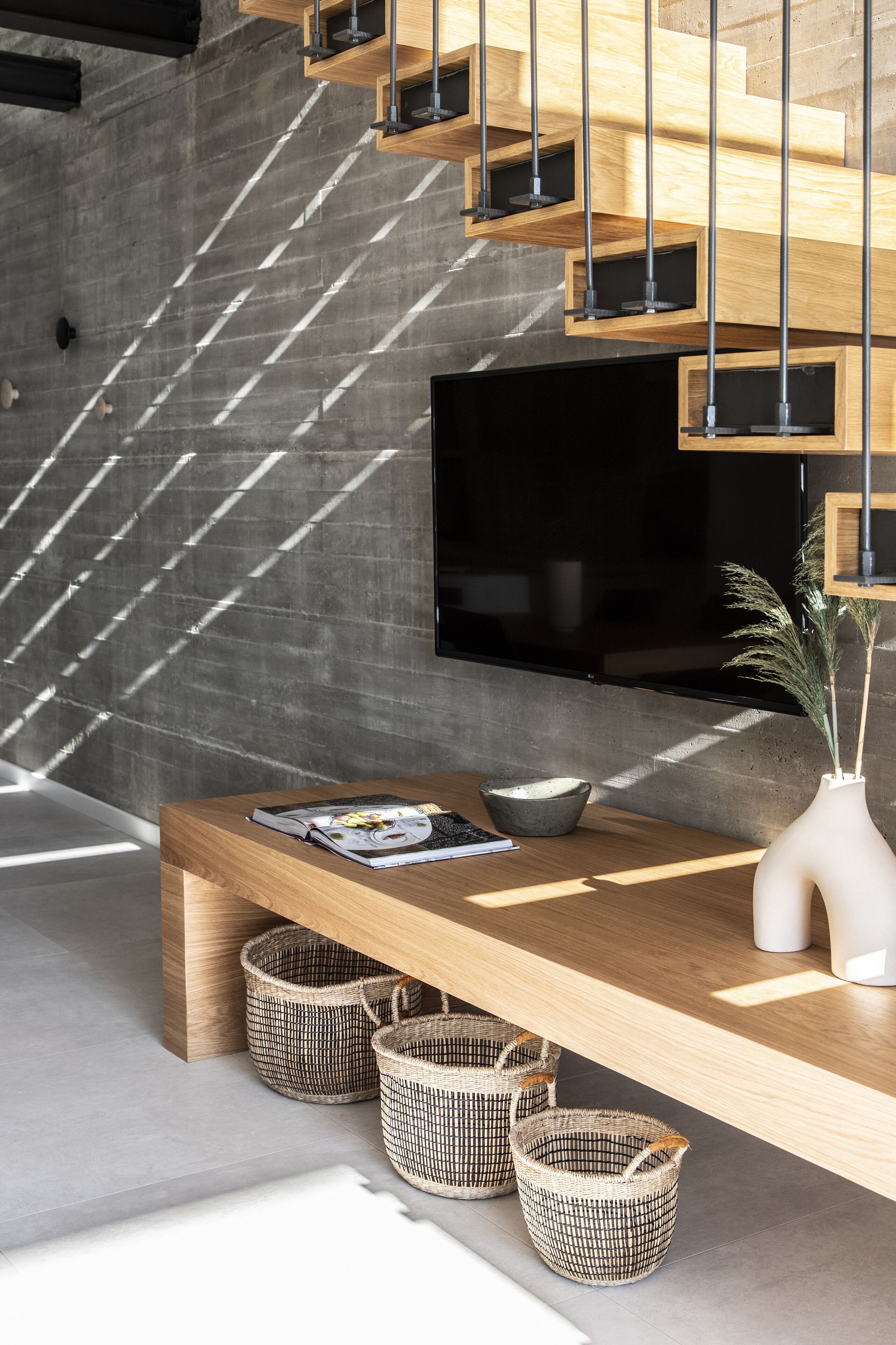 A modern interior with a wooden staircase, a flat-screen TV, a wooden table with magazines, and wicker baskets.
